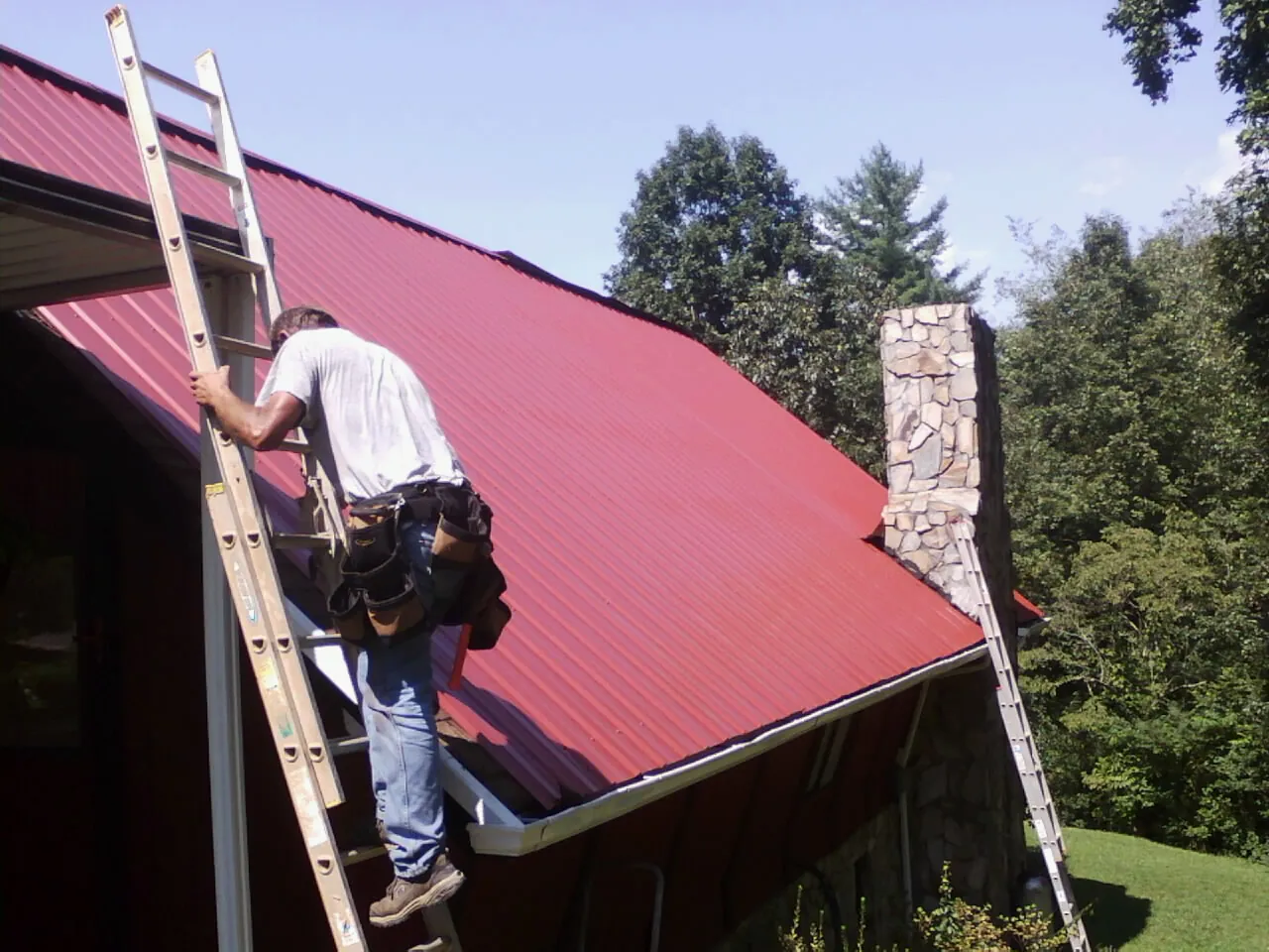 Roofer on a ladder beside a red metal roof and stone chimney — Soffit & Fascia Repair by Roofing Brothers in Ramsay